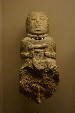 A color photo of the bard with lyre buste. It is suspended on a wall by a wire that wraps beneath His torc. He has simple, almond shaped eyes with a tall triangular nose and a single slit for a mouth. He has stylized curly ears. He holds a large seven-string lyre in front of himself with both hands. The lyre has a sound box and its curved arms curl at the top where they join the crossbar.