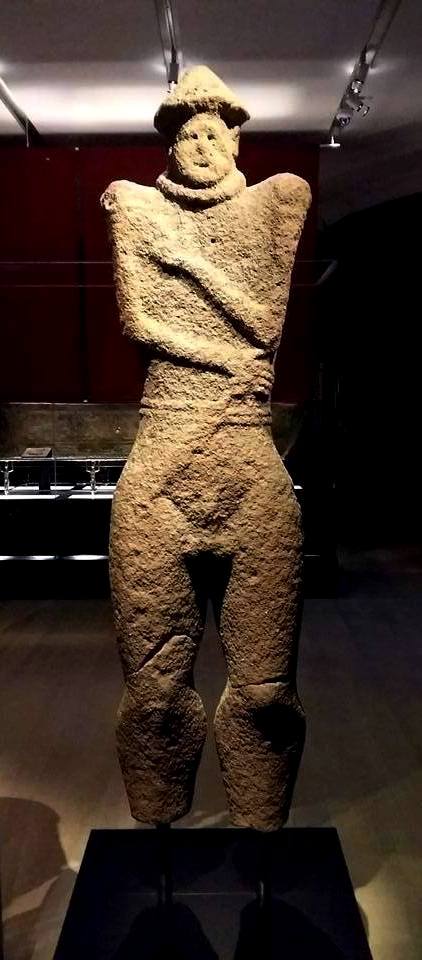 Photo of a heavily worn, beige stone statue of a man, under a museum spotlight. He wears a conical hat and has sharp shoulders that jut upwards like he's shrugging. His face is simple dots for eyes and line for a mouth. He has a collar around his neck (maybe a torque) and His left hand folds over His chest while His right hand holds a dagger towards the floor. His legs are massive like tree trunks. Ityphallic genitalia can faintly be discerned