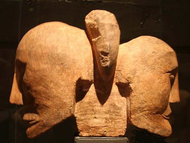 Two brown, stone heads attached to a rectangular plinth facing opposite directions in the style of Janus. There is an oblong stone in between the tops of the heads, perhaps like one of the so-called misteltoe leaves. The heads have stark, angular features chiseled into them, with long, somber expressions on their faces.